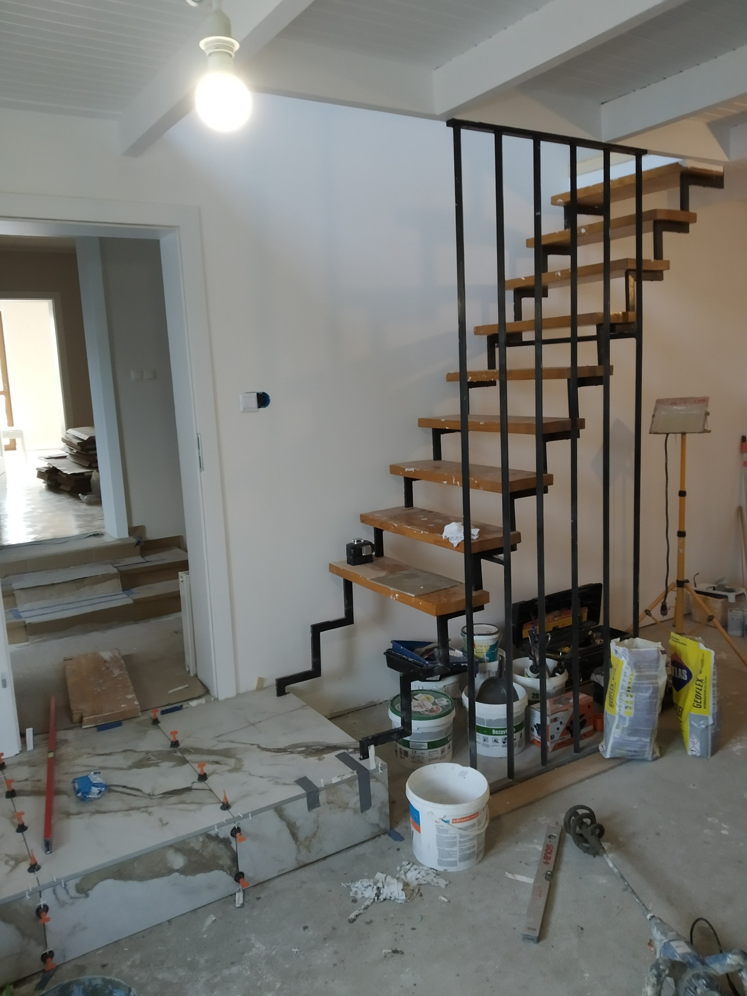 Montaż ażurowych schodów z drewnianymi stopniami i metalową konstrukcją w trakcie remontu, widoczne narzędzia i materiały budowlane, płytki podłogowe w trakcie układania.