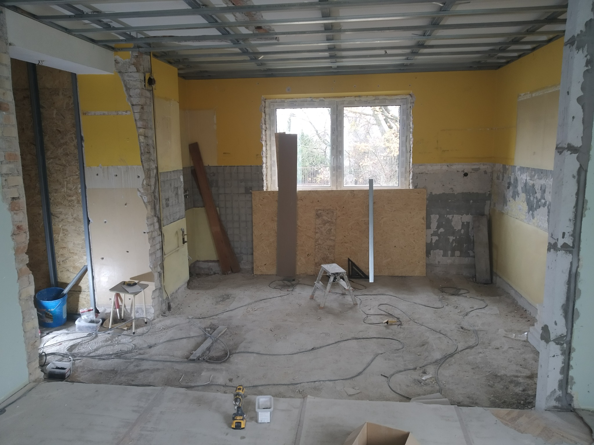 Wnętrze pokoju w trakcie remontu: odsłonięte cegły, żółte ściany, metalowa konstrukcja sufitu, okno z widokiem na drzewa, narzędzia na podłodze.