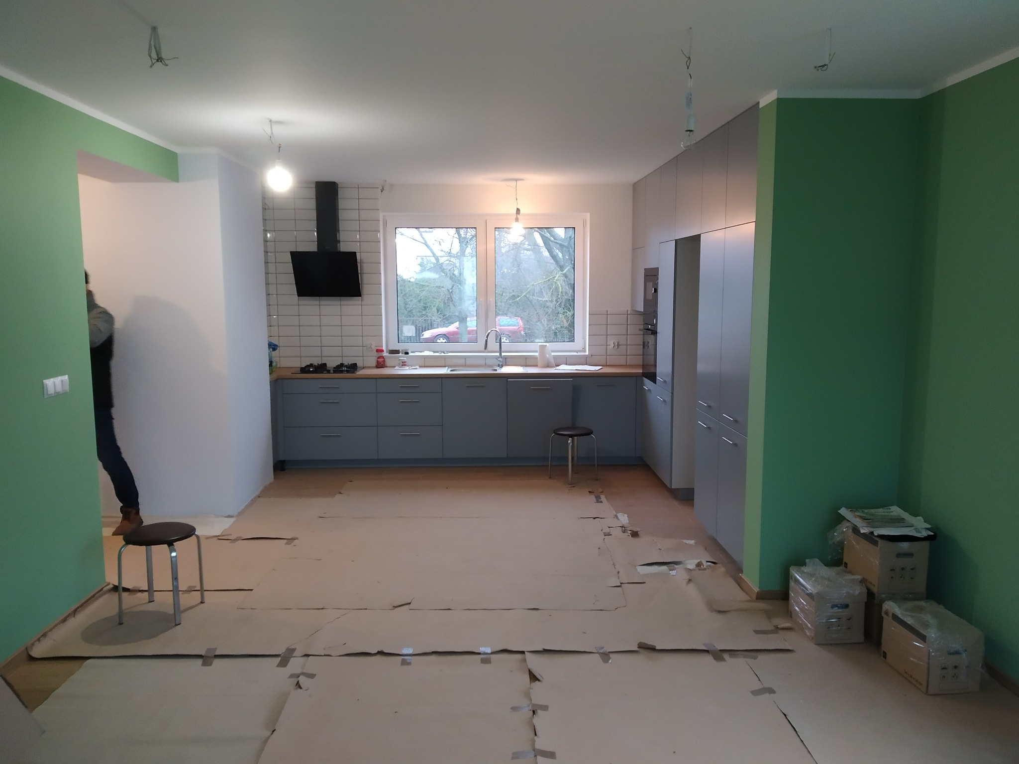 Wnętrze kuchni w trakcie remontu: szare szafki kuchenne, białe płytki na ścianie, podłoga zabezpieczona kartonem, ściany pomalowane na zielono, widoczne przygotowania do dalszych prac wykończeniowych.