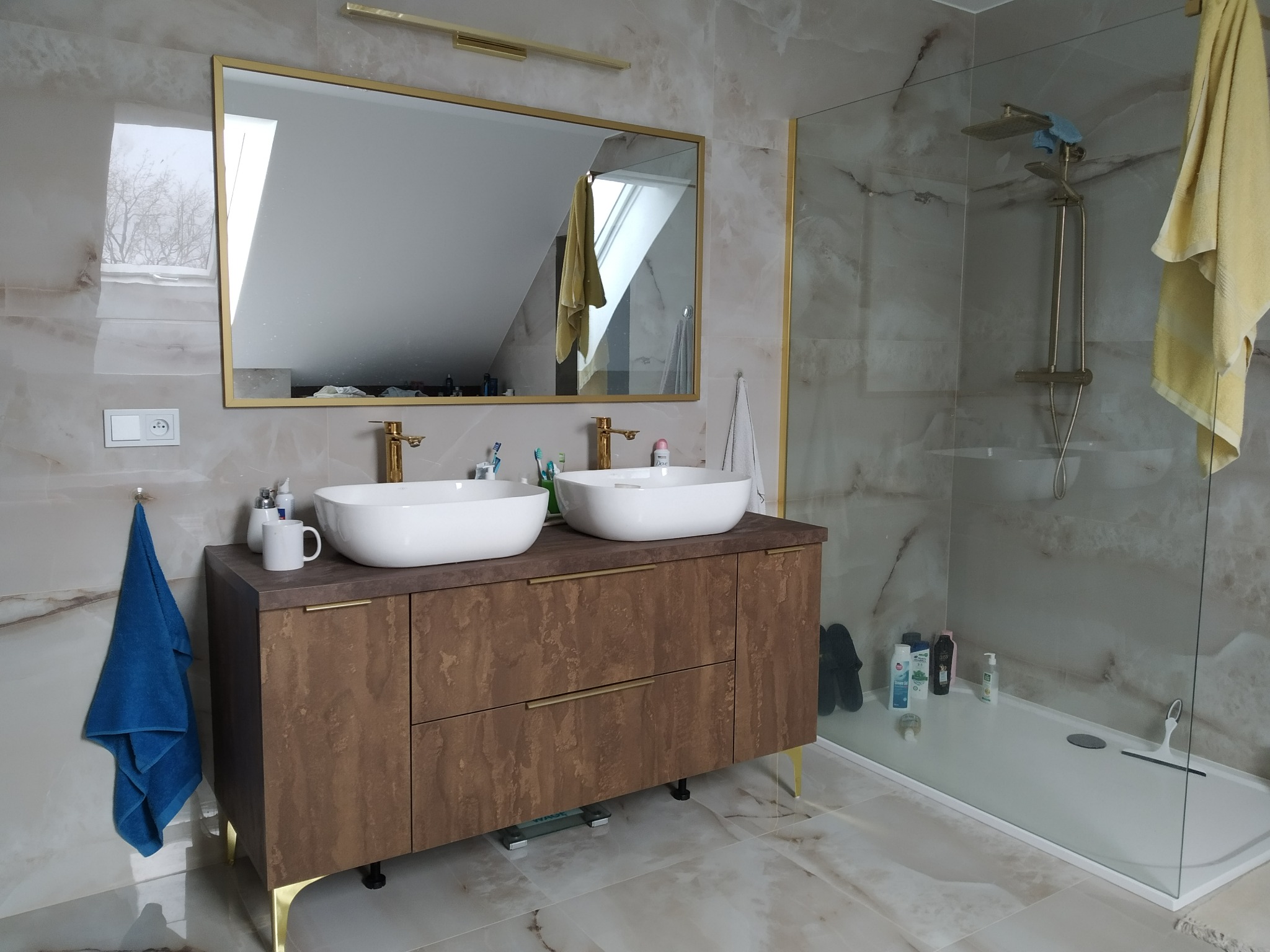 Nowoczesna łazienka z dwoma umywalkami nablatowymi, brązową szafką z złotymi nóżkami i armaturą, kabiną prysznicową ze szklaną ścianą i marmurowymi płytkami.