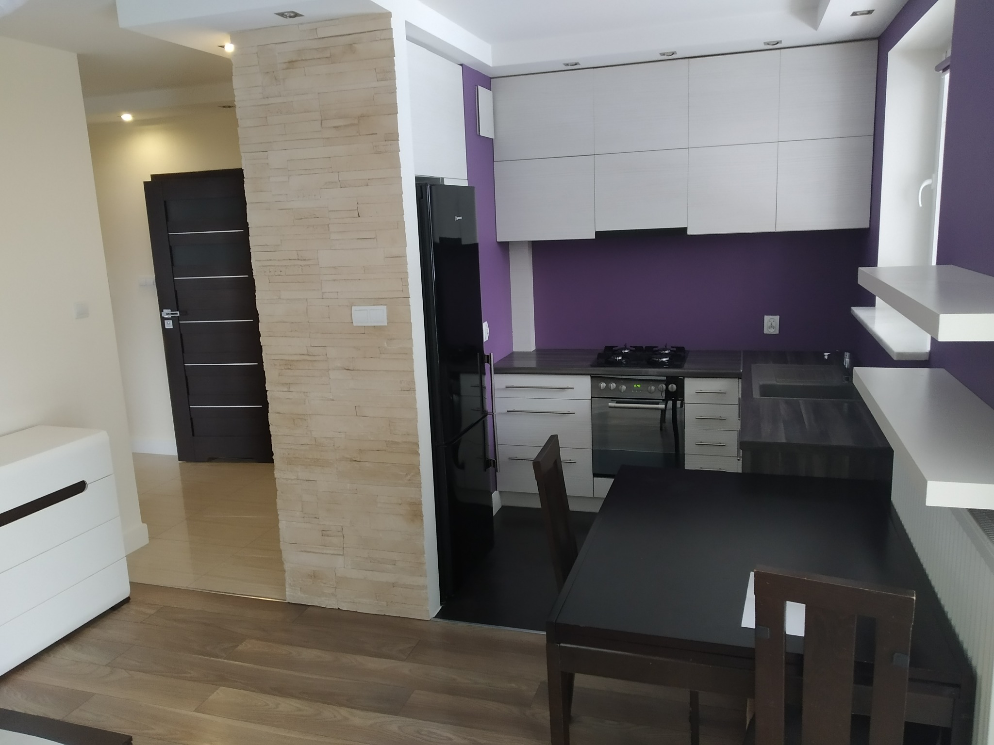 Nowoczesna kuchnia z białymi szafkami, fioletową ścianą, czarnym blatem i stołem z krzesłami, oddzielona od salonu ścianą z dekoracyjnej cegły.