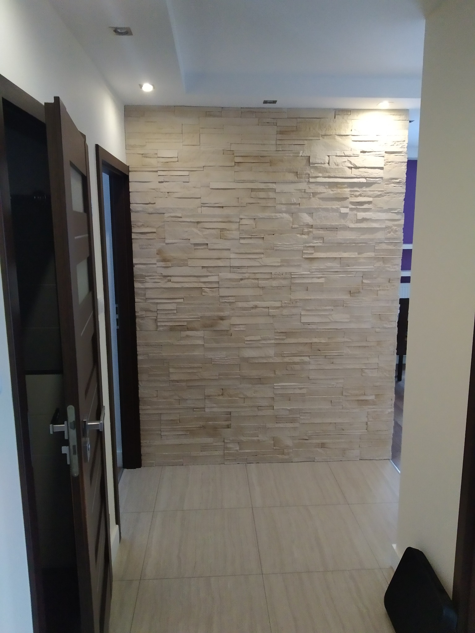 Korytarz z dekoracyjną ścianą z paneli imitujących kamień, jasne płytki podłogowe, widoczne drzwi z ciemnego drewna i oświetlenie punktowe sufitowe.