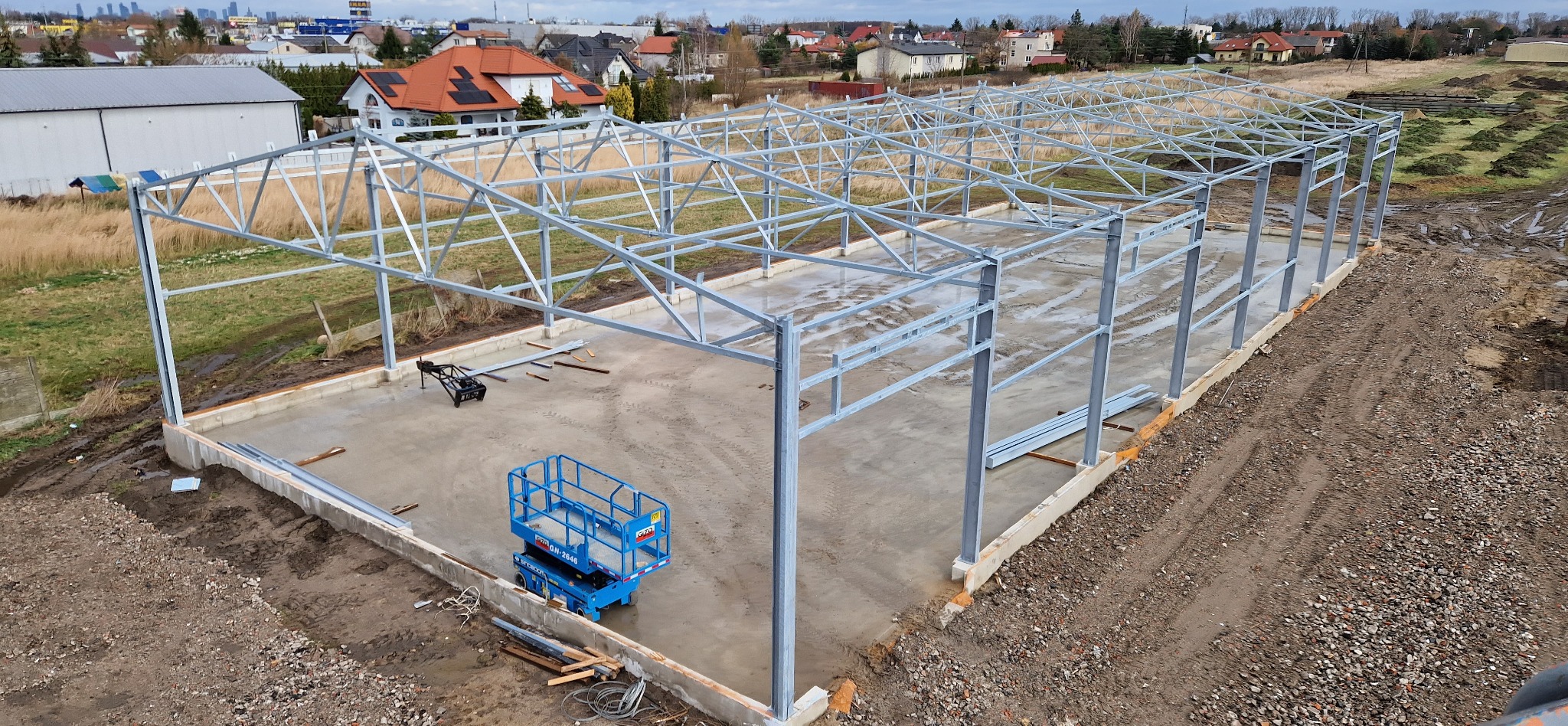 Stalowa konstrukcja hali w trakcie budowy, widoczny betonowy fundament, podnośnik koszowy i rozległy teren budowy z zabudową w tle.