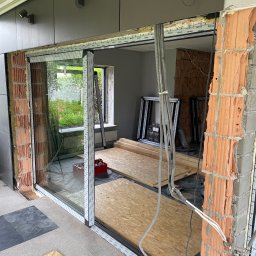 Alumaster Bud - Montaż przesuwnych okien aluminiowych z widoczną konstrukcją ściany z cegły dziurawki i elementami instalacji elektrycznej, widok na ogród przez szybę, w tle profile okienne.