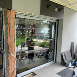 Montaż dużego okna tarasowego z użyciem przyssawek, widoczne niedokończone wykończenie muru i elementy instalacji elektrycznej.