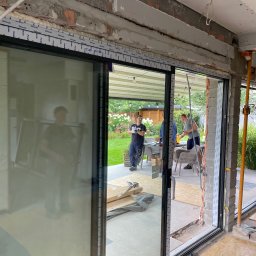 KLAMAT SPÓŁKA Z OGRANICZONĄ ODPOWIEDZIALNOŚCIĄ - Montaż przesuwnych okien aluminiowych z widokiem na ogród i pracujących monterów, widoczne surowe wykończenie ściany i elementy montażowe.