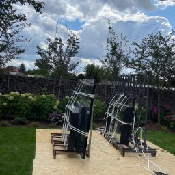 KLAMAT SPÓŁKA Z OGRANICZONĄ ODPOWIEDZIALNOŚCIĄ - Szyby okienne zabezpieczone taśmą i umieszczone na drewnianych stojakach, przygotowane do montażu na trawniku przed domem z kamiennym ogrodzeniem i kwitnącymi hortensjami w tle.