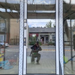 KLAMAT SPÓŁKA Z OGRANICZONĄ ODPOWIEDZIALNOŚCIĄ - Szklane drzwi wejściowe z białymi ramami, widoczne ślady montażu i odbicie osoby robiącej zdjęcie. W tle budynek z szarym dachem i roślinami.