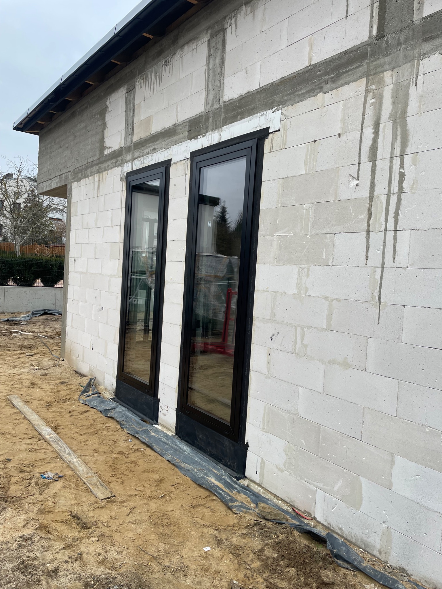 Dwa okna PCV z czarnymi ramami zamontowane w surowej ścianie z betonu komórkowego na placu budowy, widoczne ślady po zaprawie i folia ochronna przy podstawie.