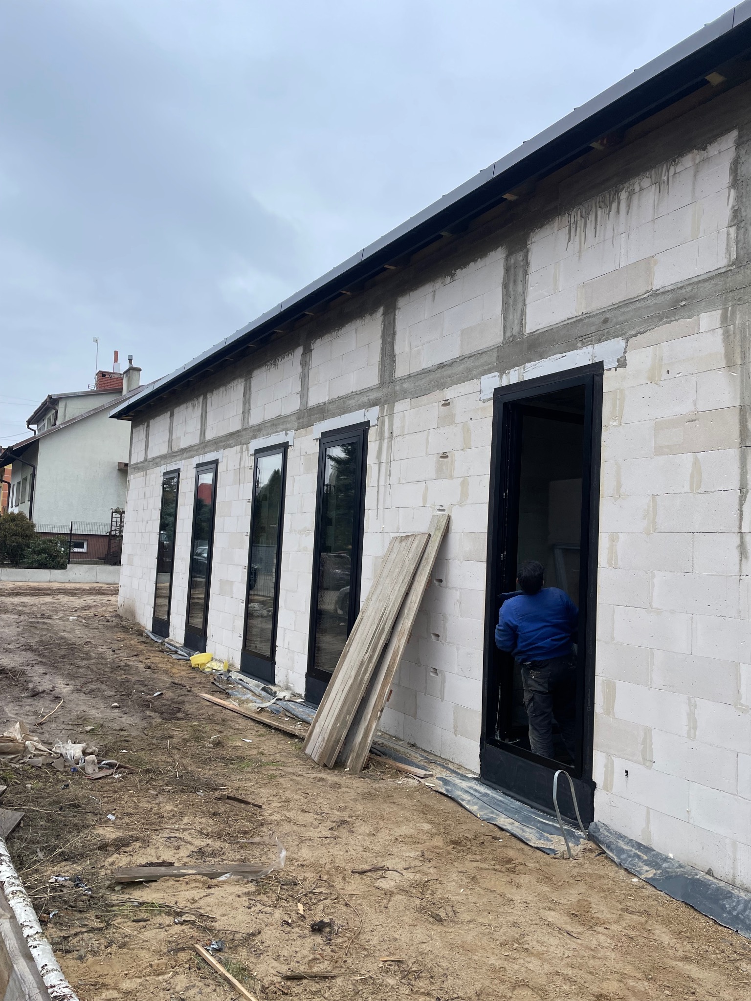 Budynek w trakcie budowy z zamontowanymi oknami i drzwiami z czarnymi ramami, widoczny pracownik w środku, na zewnątrz leżą deski.
