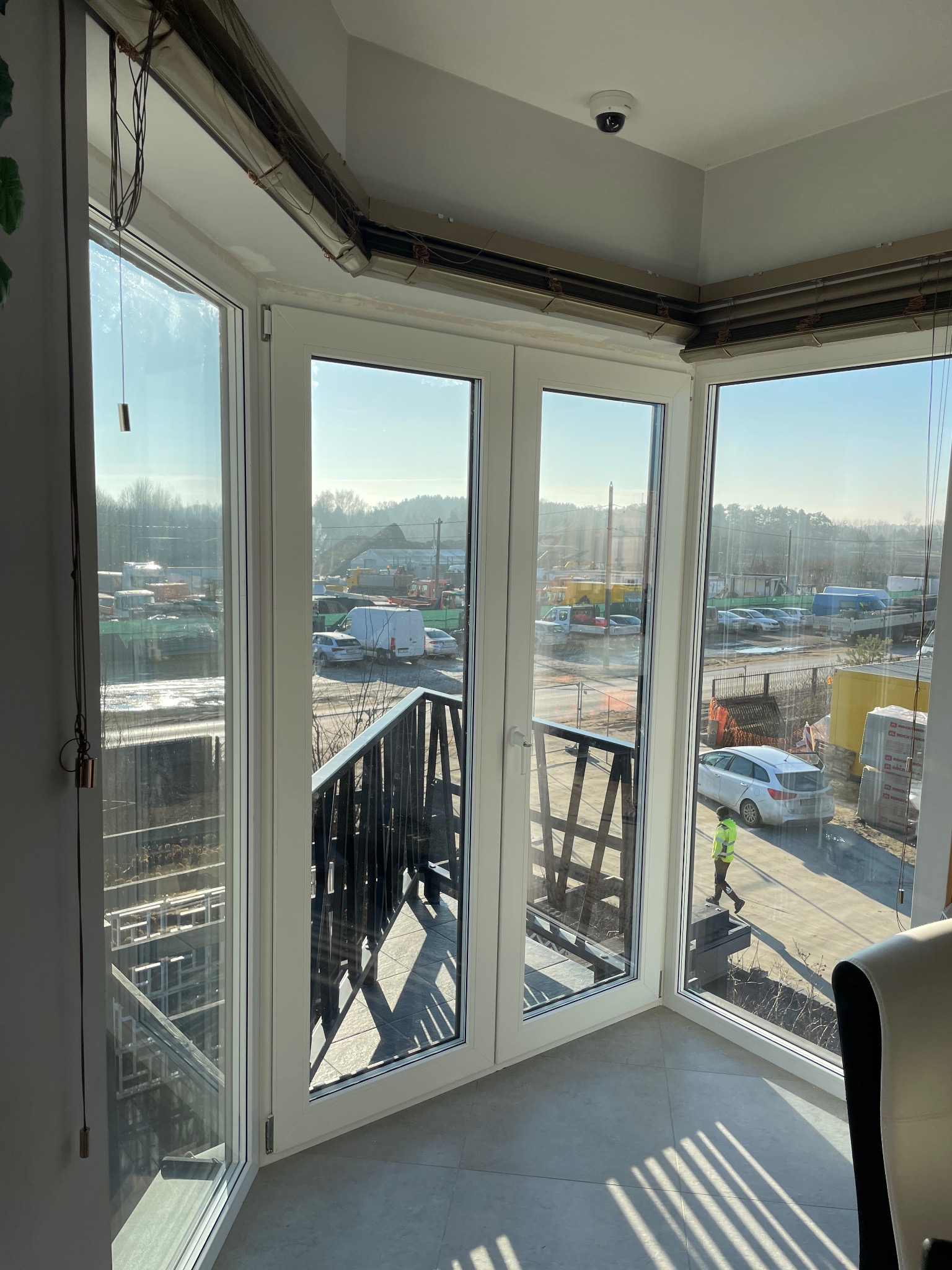 Widok z wnętrza przez przeszklone, białe drzwi balkonowe na plac budowy z widocznymi pojazdami, materiałami budowlanymi i osobą w jaskrawej kurtce. Na suficie widoczna kamera monitoringu.