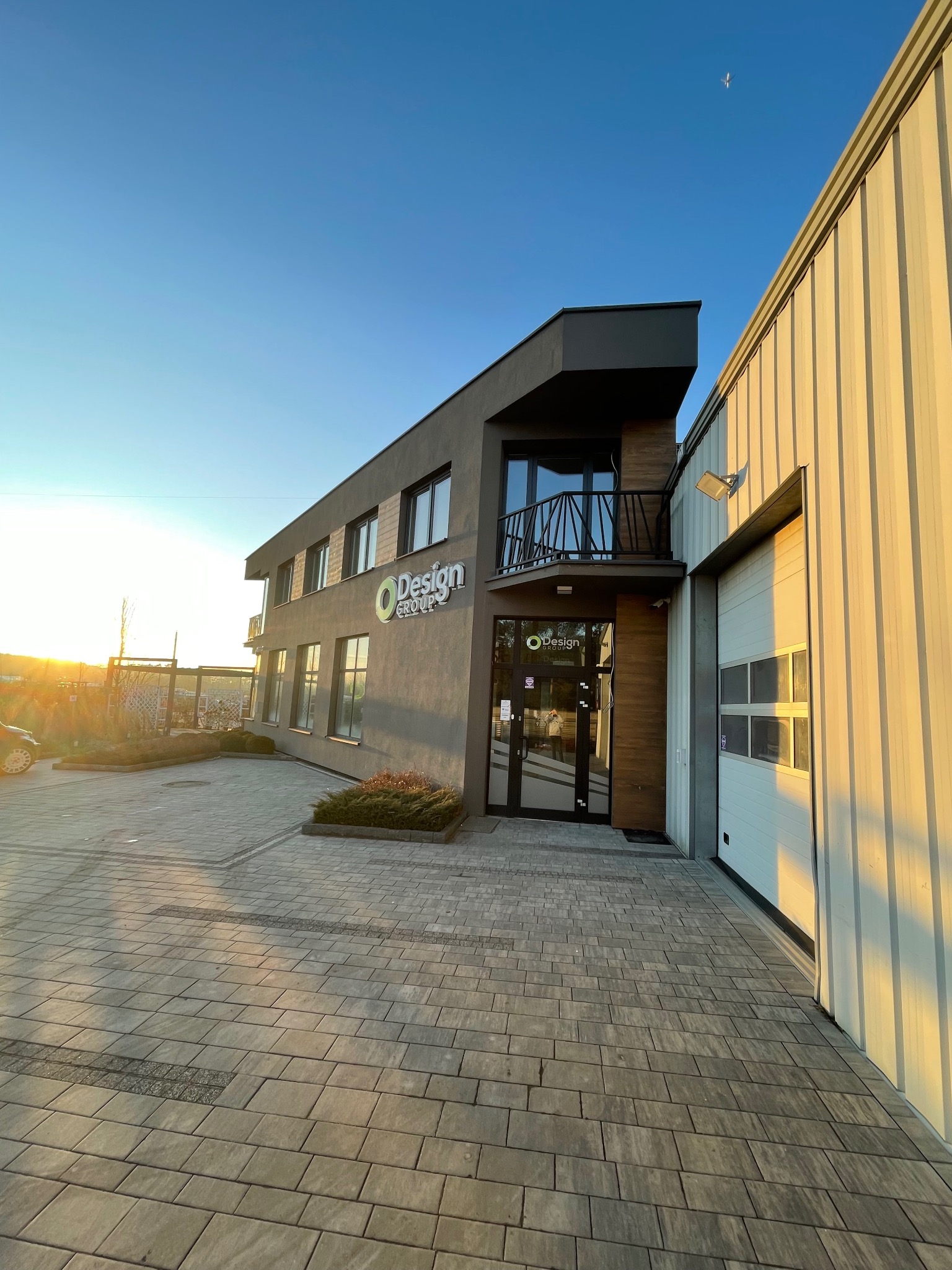 Nowoczesny budynek biurowy z elementami metalowymi, widoczne okna, drzwi wejściowe, brama garażowa i logo firmy w promieniach zachodzącego słońca.