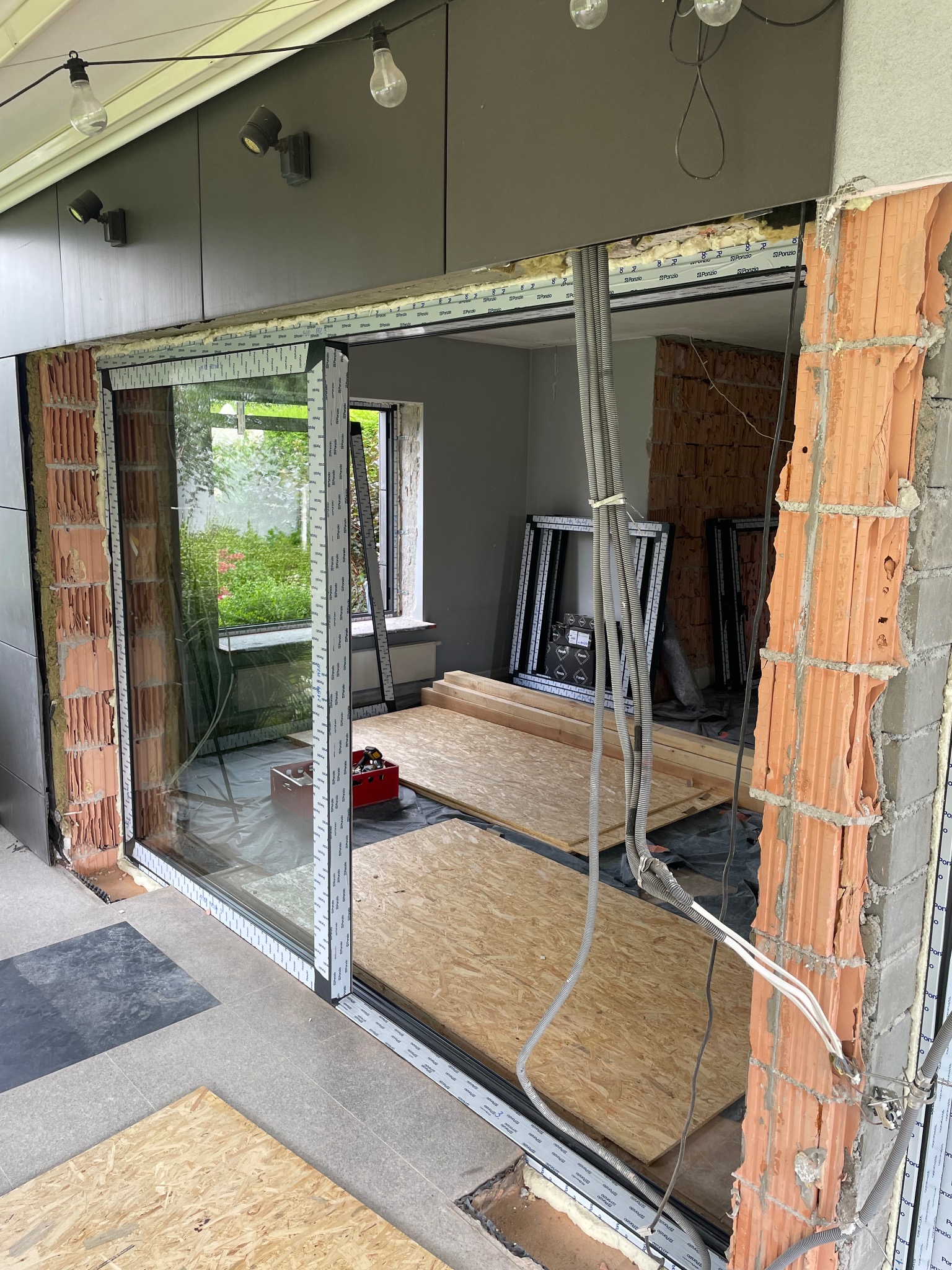 Montaż przesuwnych okien aluminiowych z widoczną konstrukcją ściany z cegły dziurawki i elementami instalacji elektrycznej, widok na ogród przez szybę, w tle profile okienne.