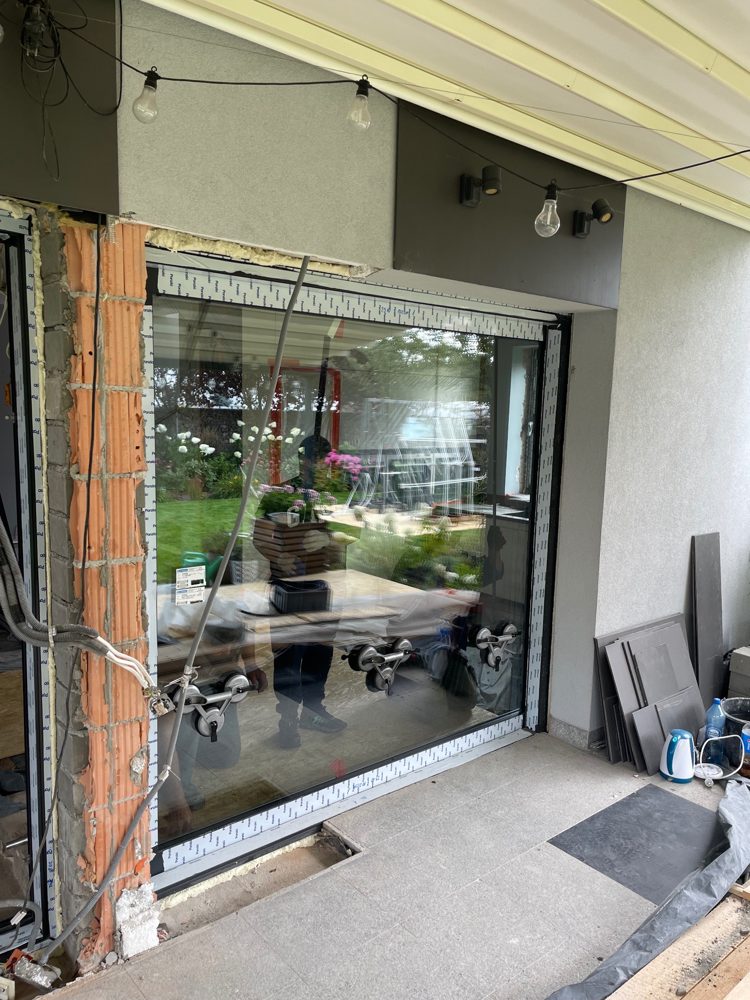 Montaż dużego okna tarasowego z użyciem przyssawek, widoczne niedokończone wykończenie muru i elementy instalacji elektrycznej.