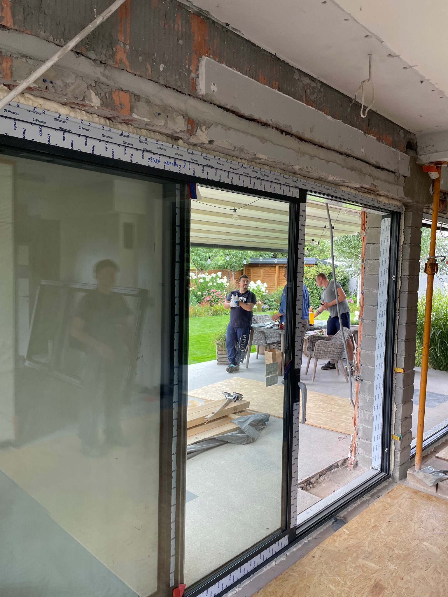 Montaż przesuwnych okien aluminiowych z widokiem na ogród i pracujących monterów, widoczne surowe wykończenie ściany i elementy montażowe.