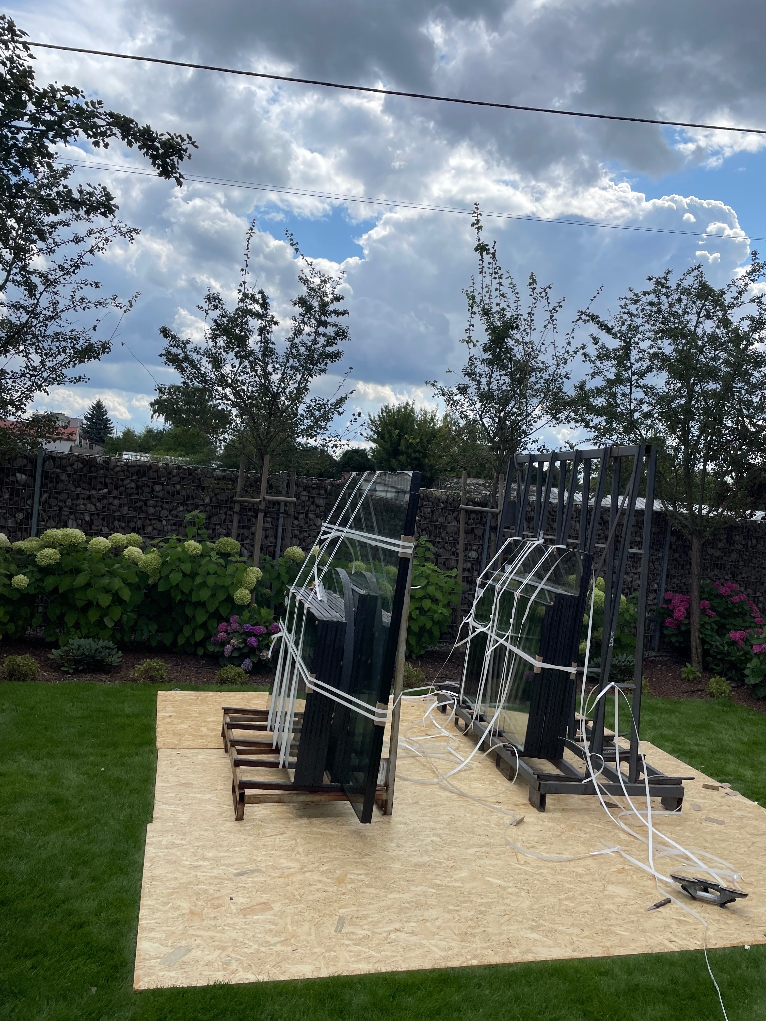 Szyby okienne zabezpieczone taśmą i umieszczone na drewnianych stojakach, przygotowane do montażu na trawniku przed domem z kamiennym ogrodzeniem i kwitnącymi hortensjami w tle.