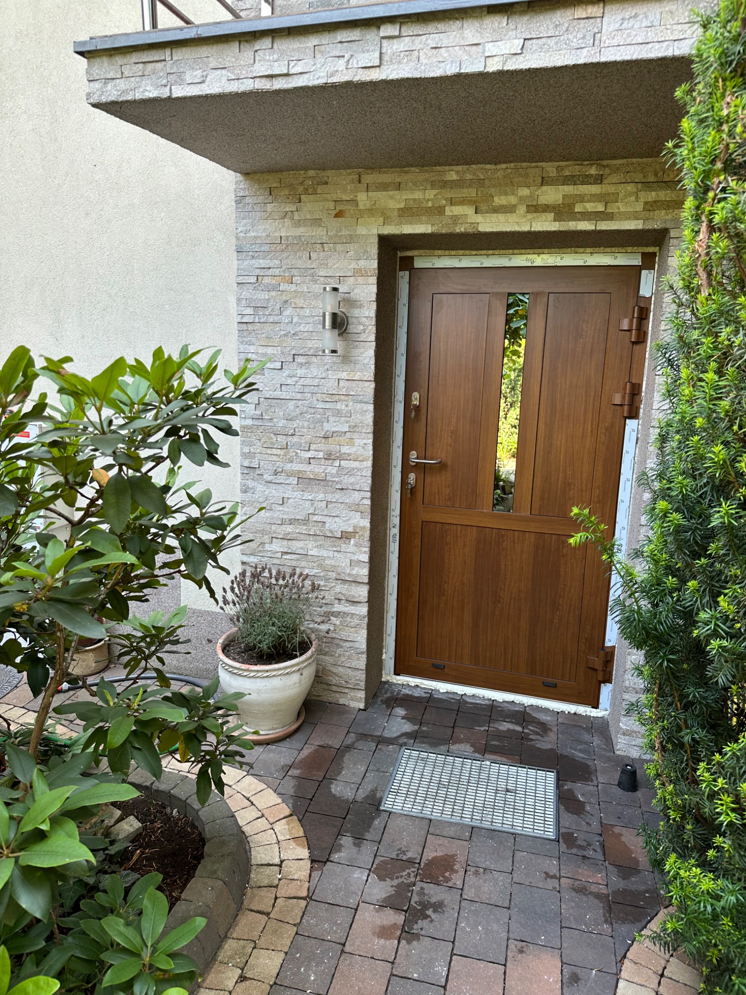 Brązowe drzwi wejściowe z przeszkleniami, osadzone w kamiennej elewacji domu, otoczone zielenią. Widoczne detale wykończenia i oświetlenie zewnętrzne.
