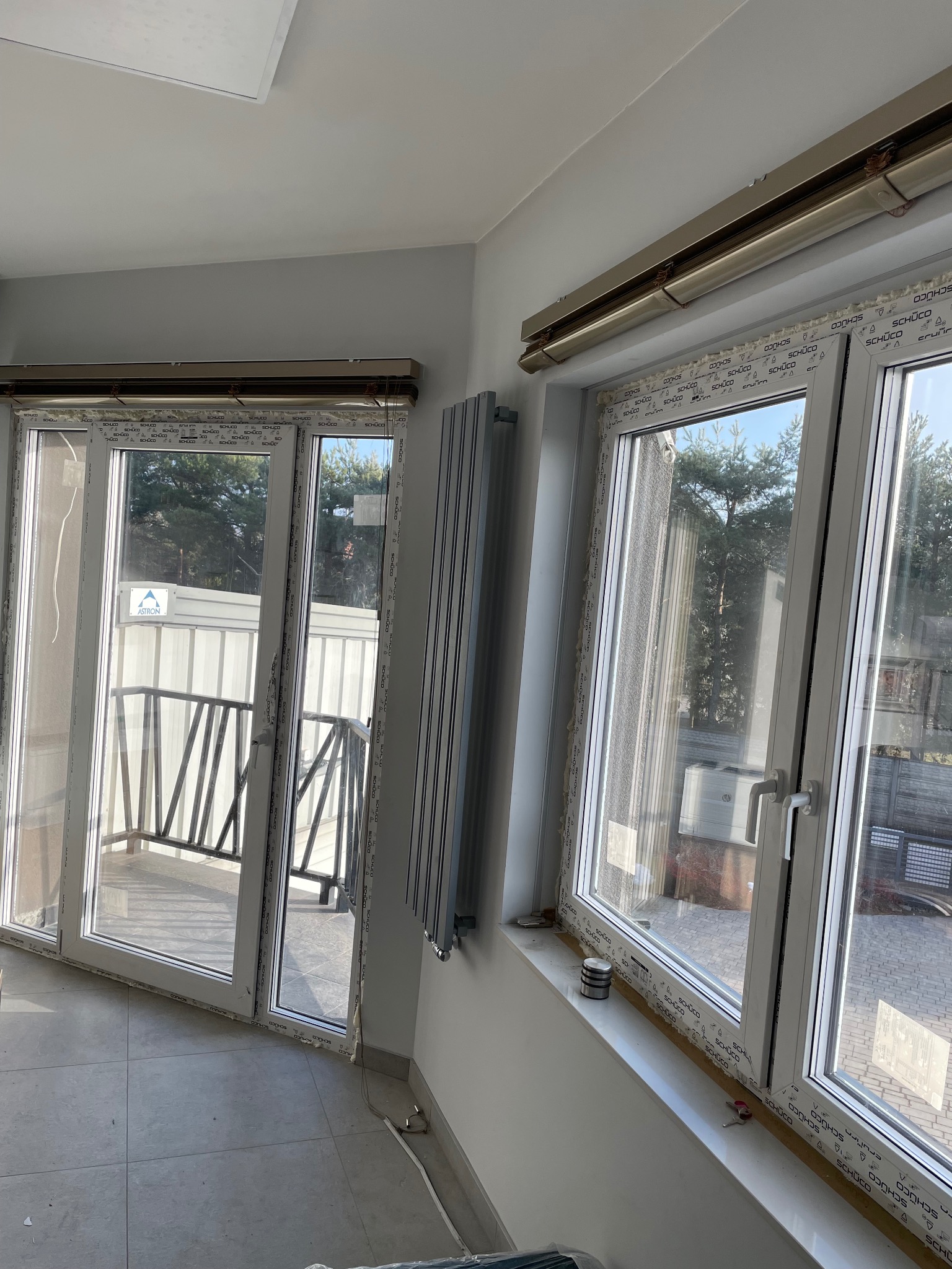 Jasne wnętrze z białymi oknami PCV marki Schüco, grzejnikiem i wyjściem na balkon. Widoczne uszczelnienia okienne i elementy montażowe. Słoneczny dzień.