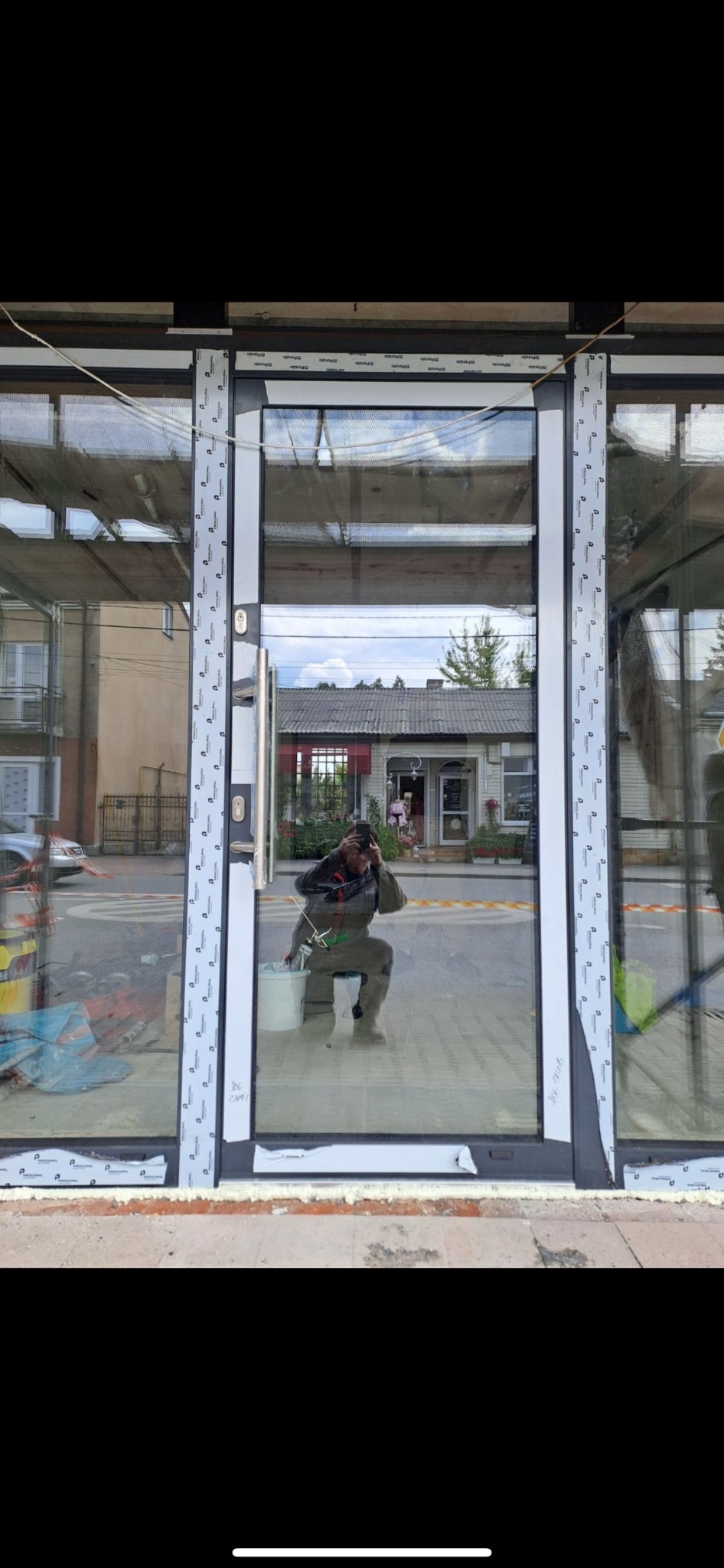 Szklane drzwi wejściowe z białymi ramami, widoczne ślady montażu i odbicie osoby robiącej zdjęcie. W tle budynek z szarym dachem i roślinami.