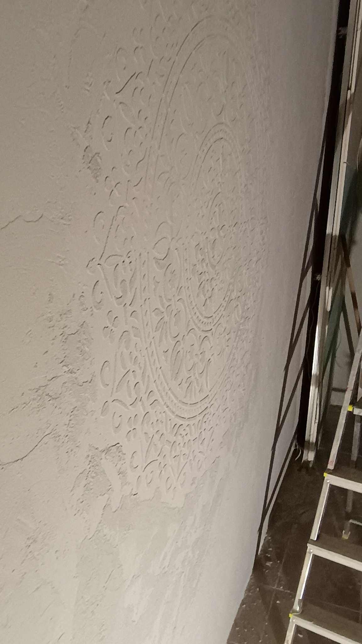 Ściana z dekoracyjnym, wypukłym wzorem mandali w odcieniach bieli, widoczna z boku, z fragmentem drabiny w prawym rogu.