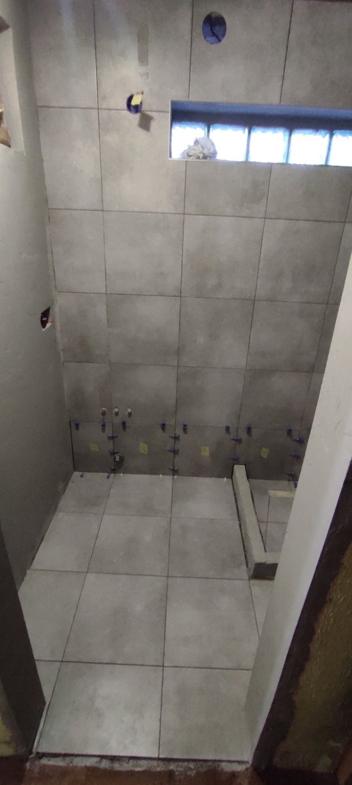 Szare płytki ceramiczne ułożone na ścianach i podłodze w surowej łazience, widoczne plastikowe krzyżyki dystansowe i gąbka w oknie.