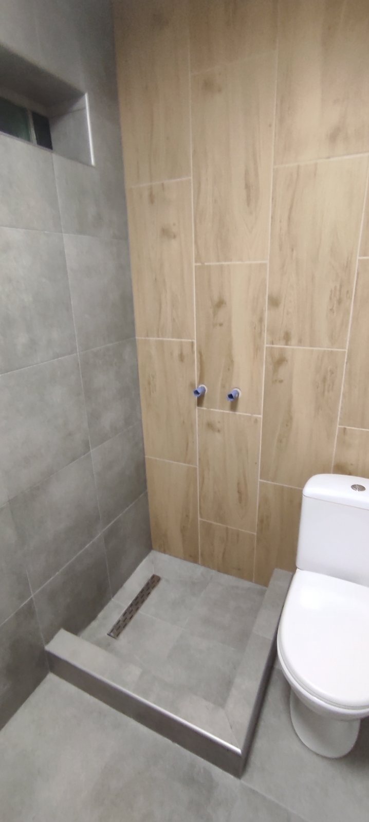 Nowoczesna łazienka z kabiną prysznicową typu walk-in wyłożoną szarymi płytkami i toaletą kompaktową, widoczne podejścia wodne na ścianie wyłożonej płytkami imitującymi drewno.