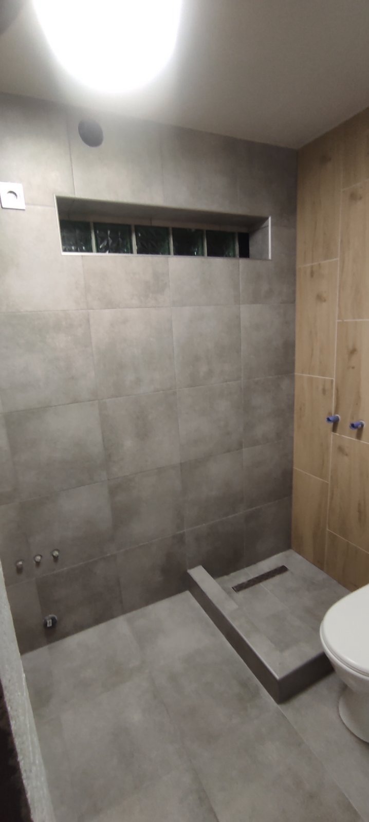 Nowoczesna łazienka z szarymi płytkami, kabina prysznicowa bez brodzika z odpływem liniowym, okno z luksferów i fragment toalety.