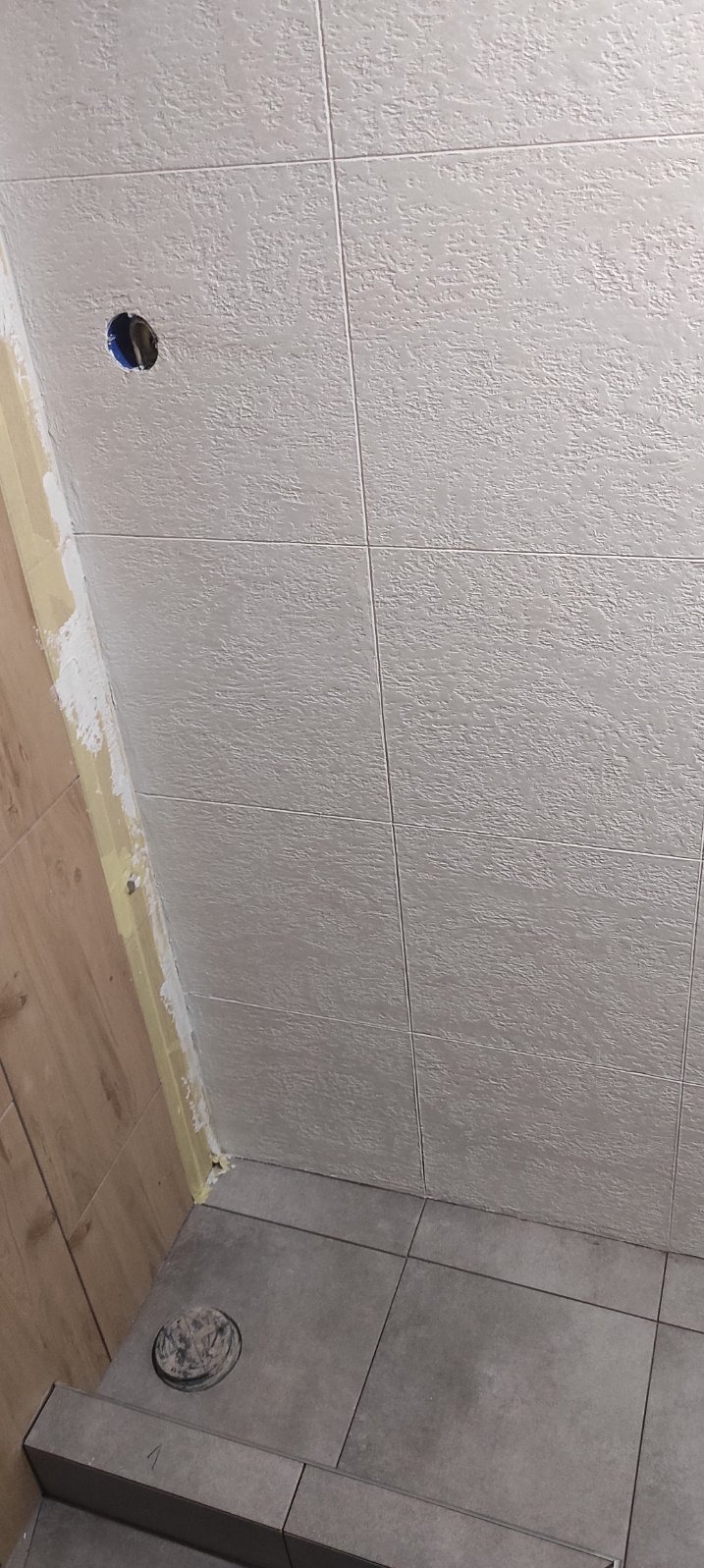 Szare płytki ścienne z fakturą i otworem na gniazdko elektryczne w trakcie montażu, widoczny fragment drewnopodobnej okładziny i szare płytki podłogowe z odpływem.