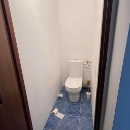 Ready Home Adrian Harabin - Wąska toaleta z białą muszlą klozetową, niebieskimi płytkami podłogowymi z wzorem marmuru i rolką papieru toaletowego leżącą na podłodze.