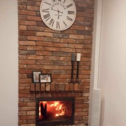 Ready Home Adrian Harabin - Ozdobny kominek z cegły z paleniskiem i płomieniami, nad nim zegar w stylu vintage i ramki ze zdjęciami, dekoracyjne świece po prawej stronie.