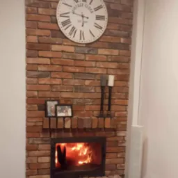 Ozdobny kominek z cegły z paleniskiem i płomieniami, nad nim zegar w stylu vintage i ramki ze zdjęciami, dekoracyjne świece po prawej stronie.