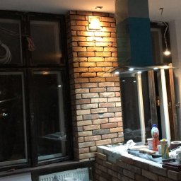 Ready Home Adrian Harabin - Wnętrze w trakcie prac: murowana obudowa okna i fragment ściany wykończone cegłą klinkierową, widoczne narzędzia i materiały budowlane, okablowanie zwisające z sufitu i z okna.