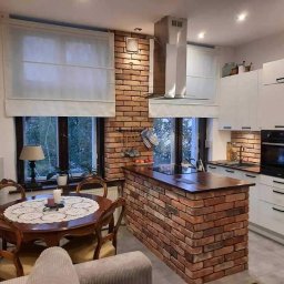 Ready Home Adrian Harabin - Kuchnia z ceglaną wyspą i ścianą, białe szafki, okna z roletami rzymskimi i drewniany stół z koronkową serwetą.