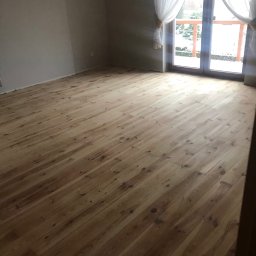 Ready Home Adrian Harabin - Jasne, drewniane deski podłogowe ułożone w pustym pokoju z dużym oknem balkonowym i białymi zasłonami, widok na zaśnieżony ogród.