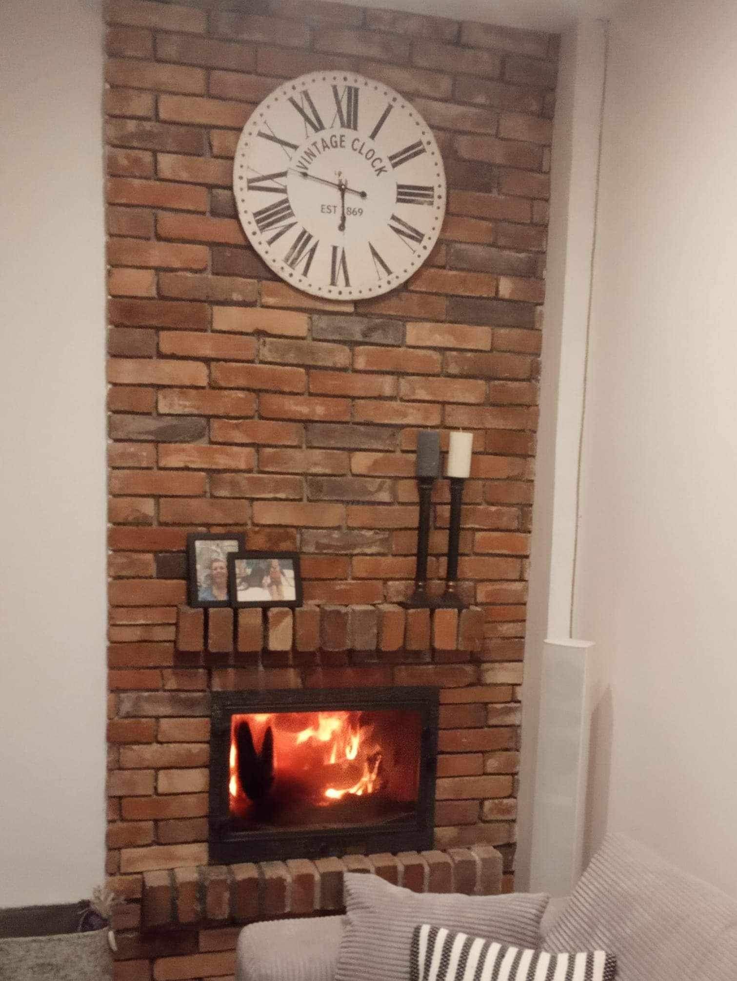 Ozdobny kominek z cegły z paleniskiem i płomieniami, nad nim zegar w stylu vintage i ramki ze zdjęciami, dekoracyjne świece po prawej stronie.