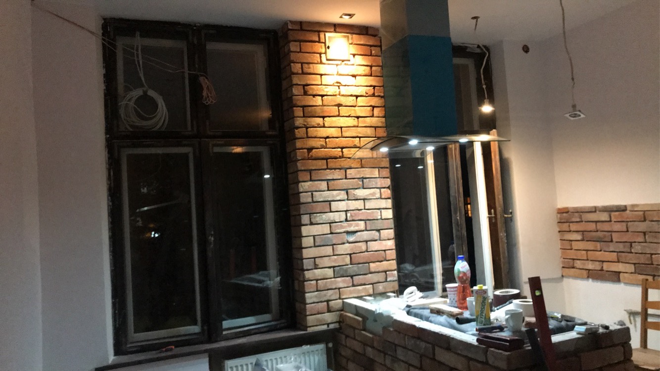 Wnętrze w trakcie prac: murowana obudowa okna i fragment ściany wykończone cegłą klinkierową, widoczne narzędzia i materiały budowlane, okablowanie zwisające z sufitu i z okna.