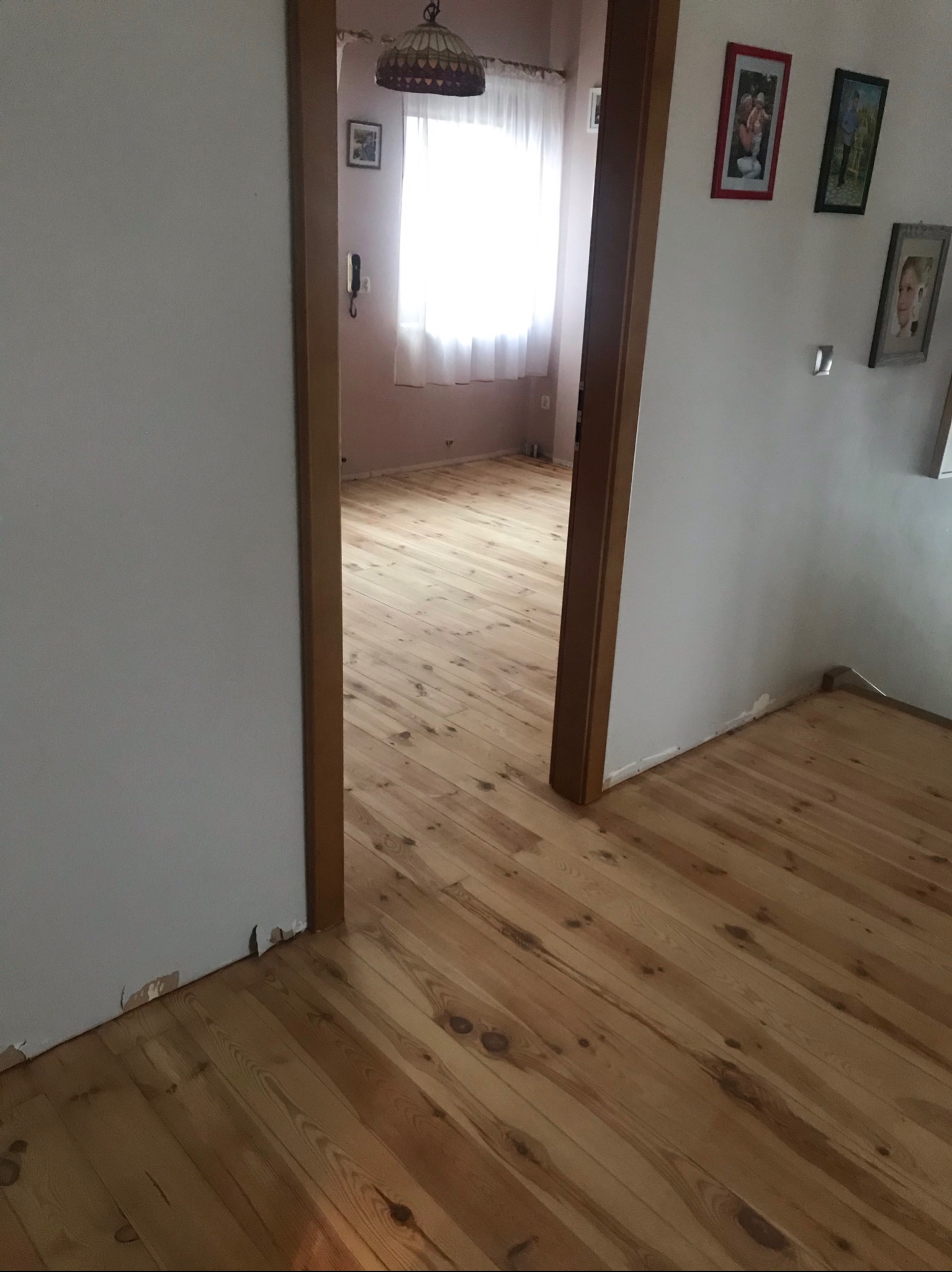 Widok na świeżo ułożoną, jasną podłogę z desek przez framugę drzwi; widoczne ślady uszkodzeń na ścianie przy podłodze.