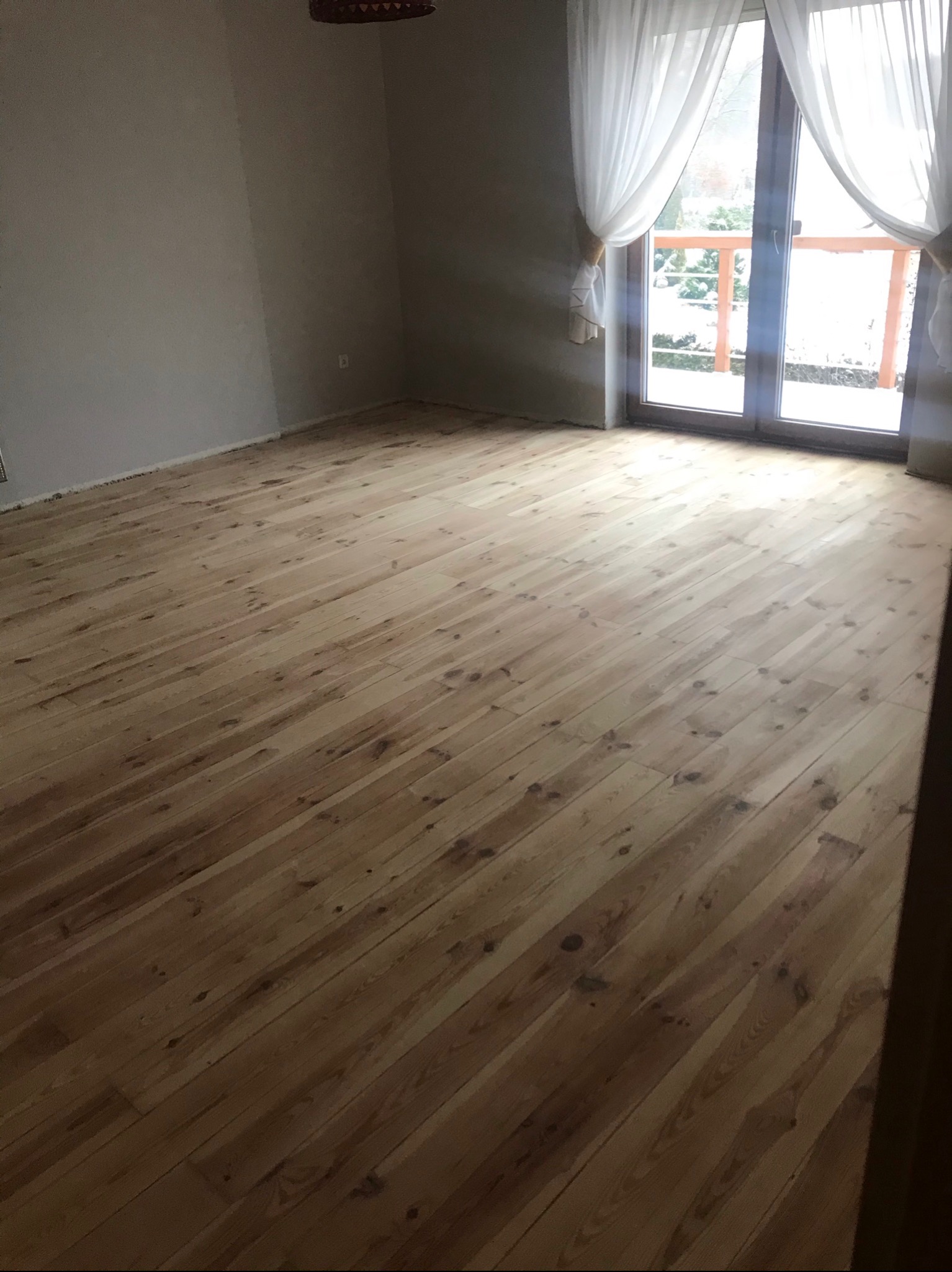 Jasne, drewniane deski podłogowe ułożone w pustym pokoju z dużym oknem balkonowym i białymi zasłonami, widok na zaśnieżony ogród.