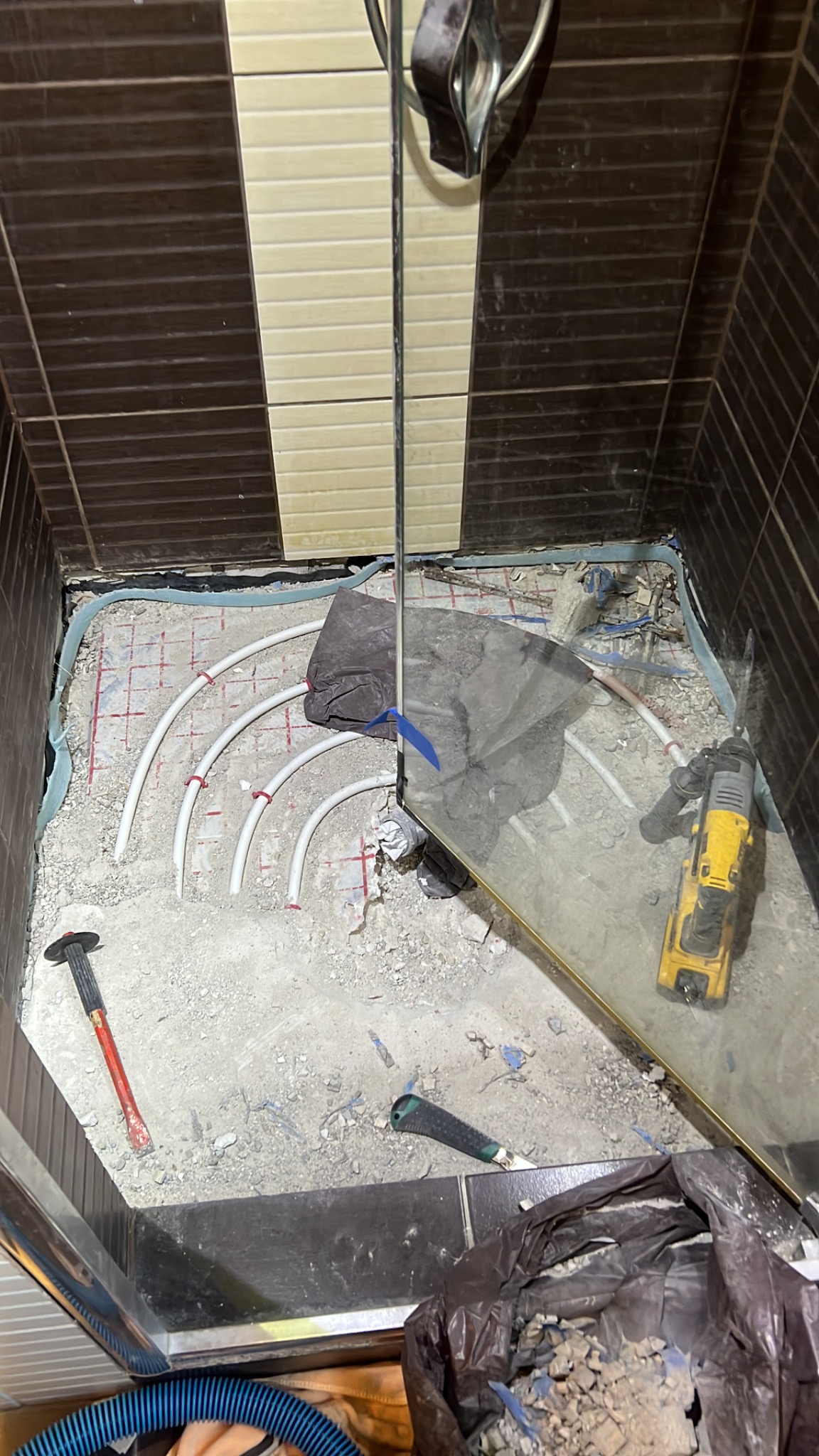 Demontaż podłogi w kabinie prysznicowej, widoczne rury ogrzewania podłogowego, gruz, narzędzia: młotek i wiertarka udarowa.