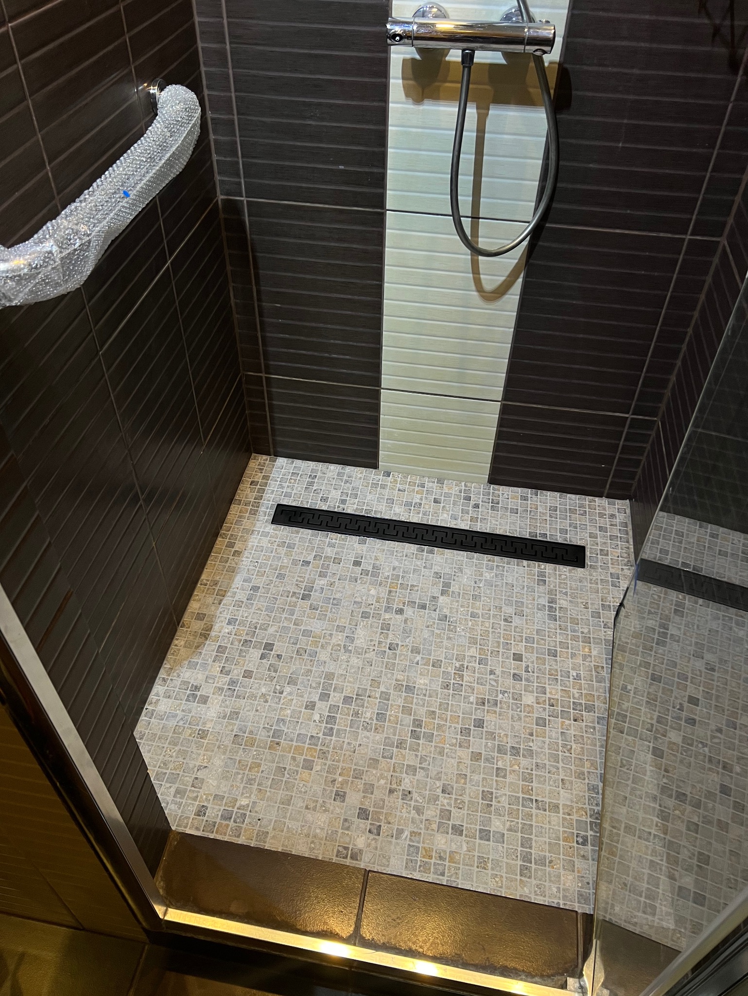 Nowoczesny brodzik z mozaikową posadzką i czarnym odpływem liniowym, ściany wyłożone brązowymi i kremowymi płytkami, chromowana bateria prysznicowa.