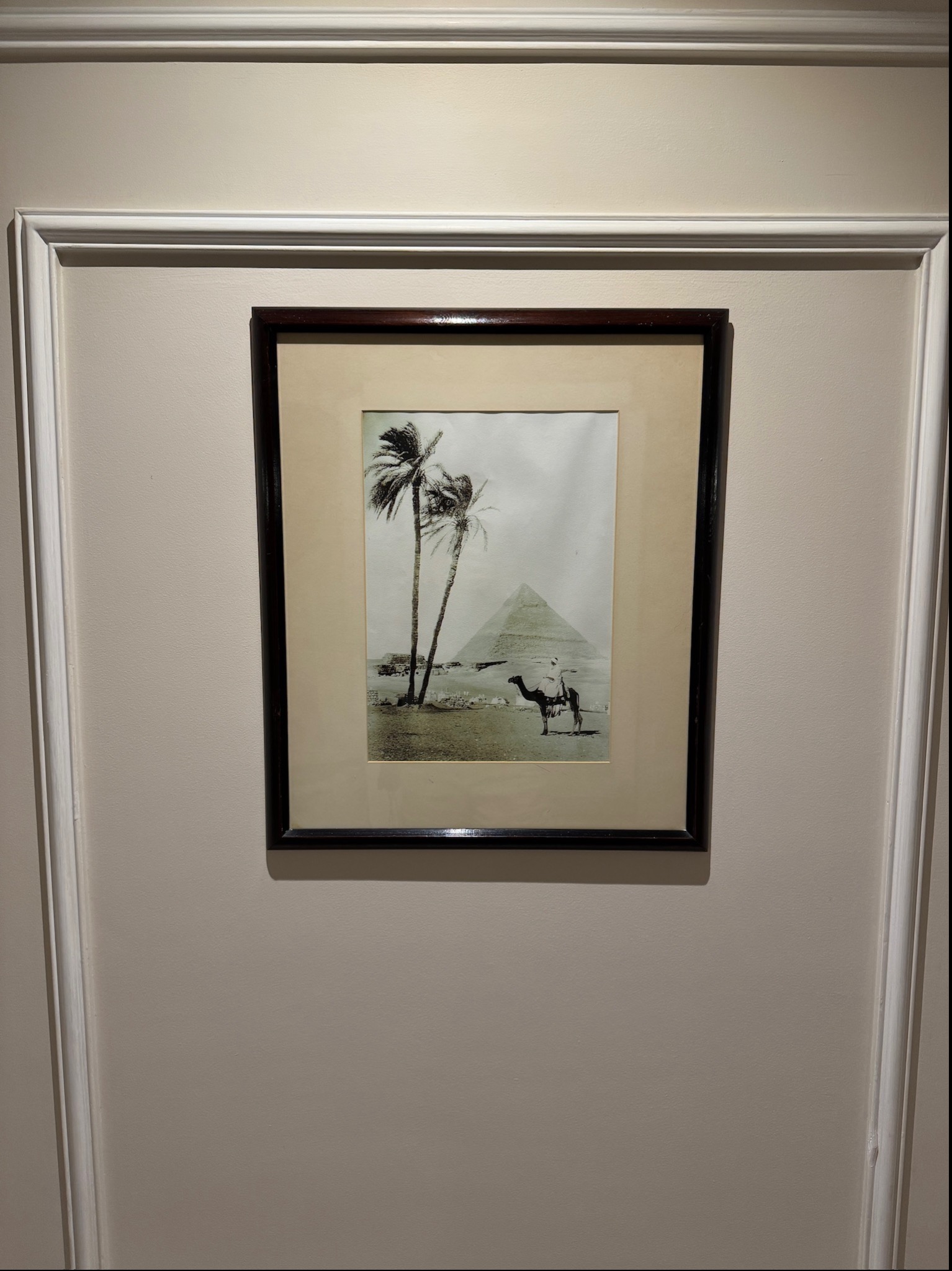 Oprawiona czarno-biała fotografia piramidy i jeźdźca na wielbłądzie, wisząca na beżowej ścianie z białą sztukaterią.