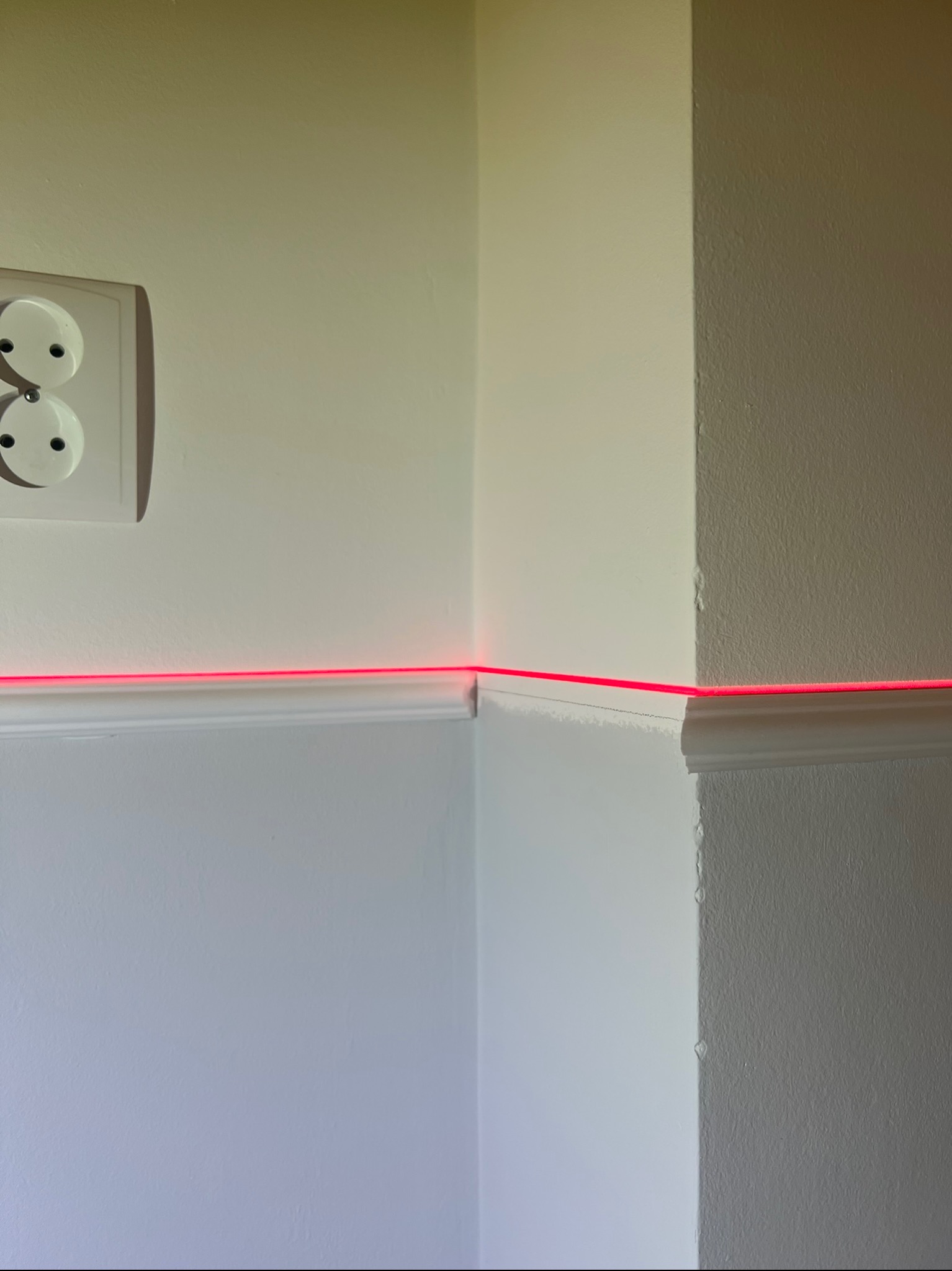 Biała ściana z listwą przypodłogową, wyznaczona laserowo czerwona linia pozioma, gniazdko elektryczne po lewej stronie.