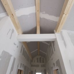 Zakład budowlany - Sufit i ściany w trakcie montażu płyt gipsowo-kartonowych, widoczne drewniane belki konstrukcyjne i szpachlowane łączenia. W tle okno.