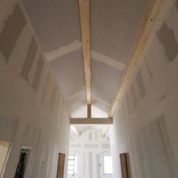 Zakład budowlany - Poddasze z płytami gipsowo-kartonowymi, widok z dołu na drewniane belki konstrukcyjne i fragment ściany z widocznymi łączeniami płyt. W tle okno i drabina.