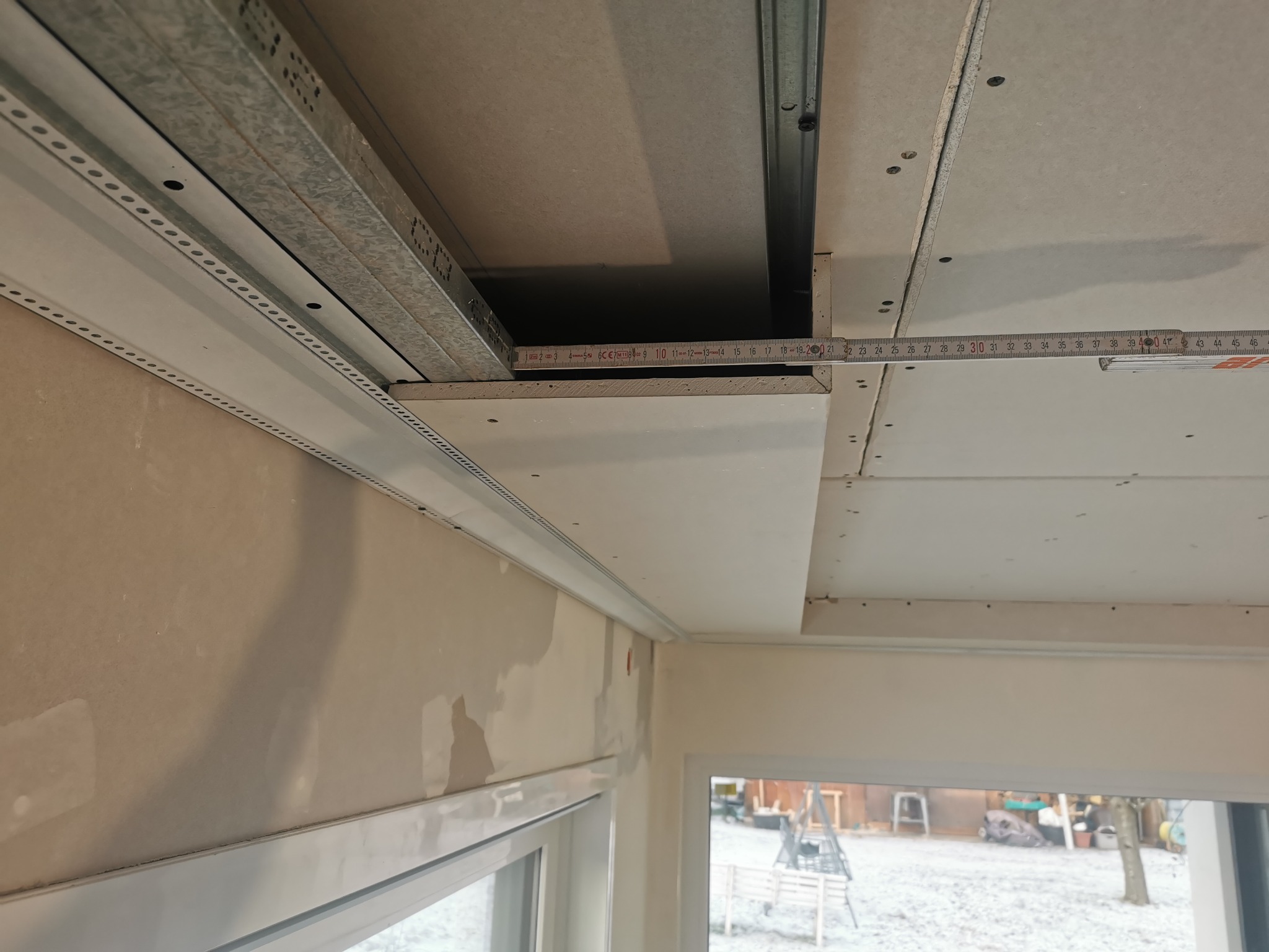 Montaż sufitu podwieszanego z płyt gipsowo-kartonowych, widoczne metalowe profile i mierząca odległość miarka. Częściowo zaszpachlowane ściany i okno z widokiem na zewnątrz.