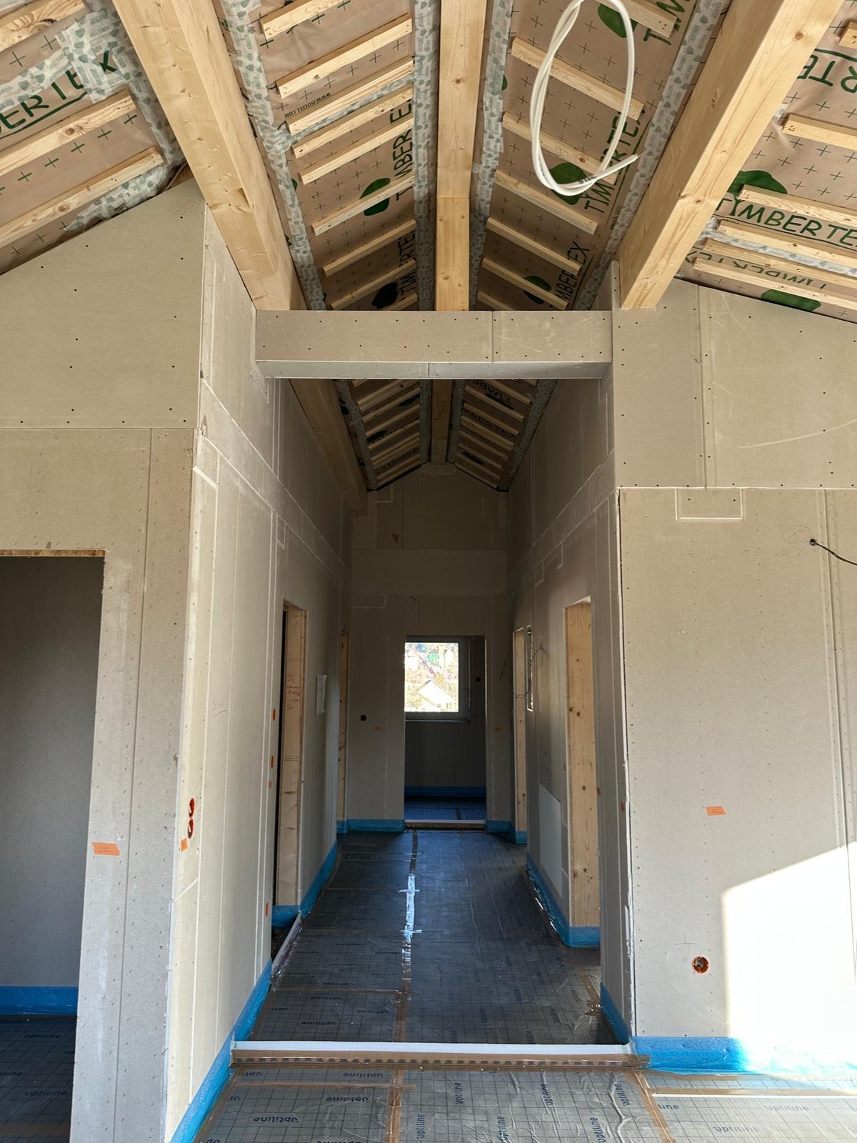 Wnętrze domu w trakcie prac wykończeniowych: widoczne płyty karton-gips na ścianach, drewniana konstrukcja dachu i folia na podłodze. Perspektywa długa, w głąb pomieszczenia.