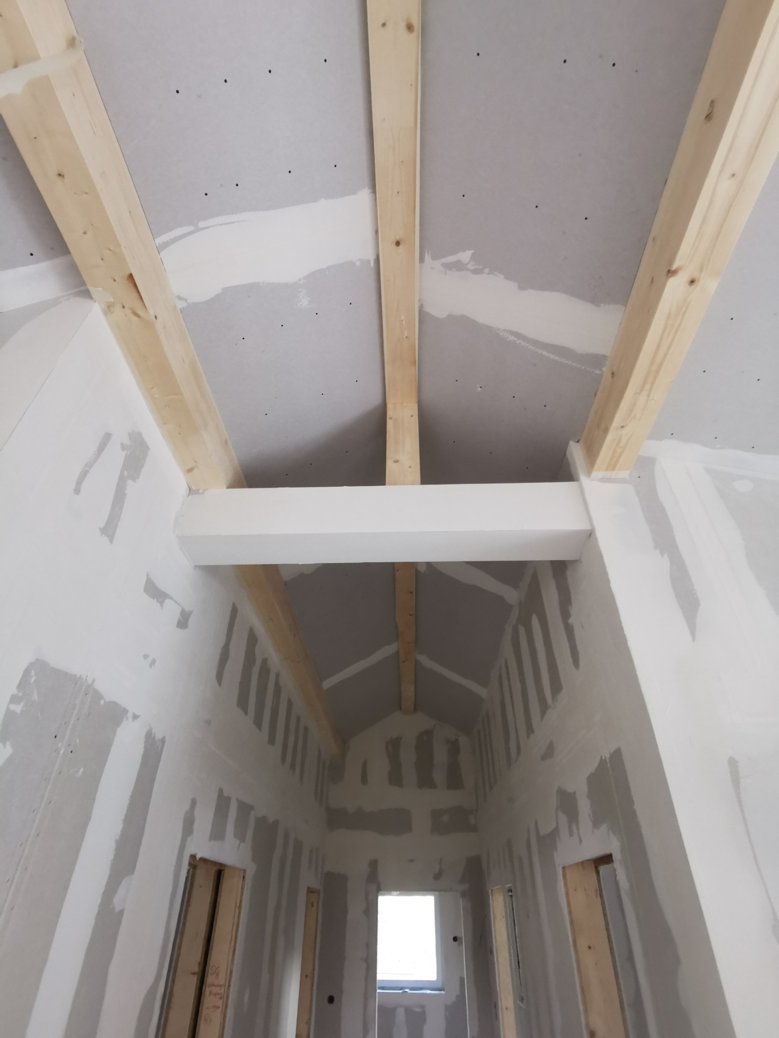 Sufit i ściany w trakcie montażu płyt gipsowo-kartonowych, widoczne drewniane belki konstrukcyjne i szpachlowane łączenia. W tle okno.