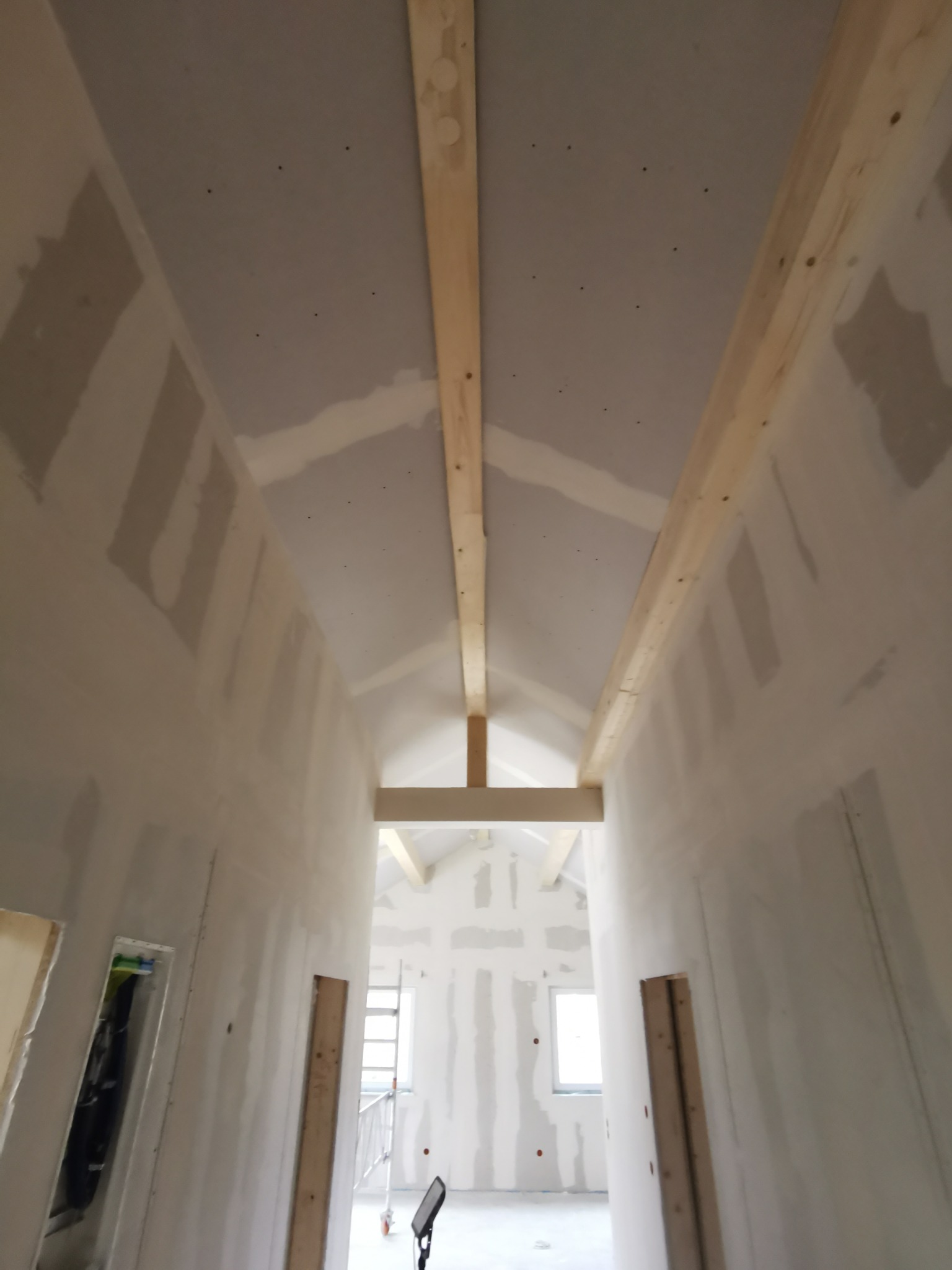 Poddasze z płytami gipsowo-kartonowymi, widok z dołu na drewniane belki konstrukcyjne i fragment ściany z widocznymi łączeniami płyt. W tle okno i drabina.