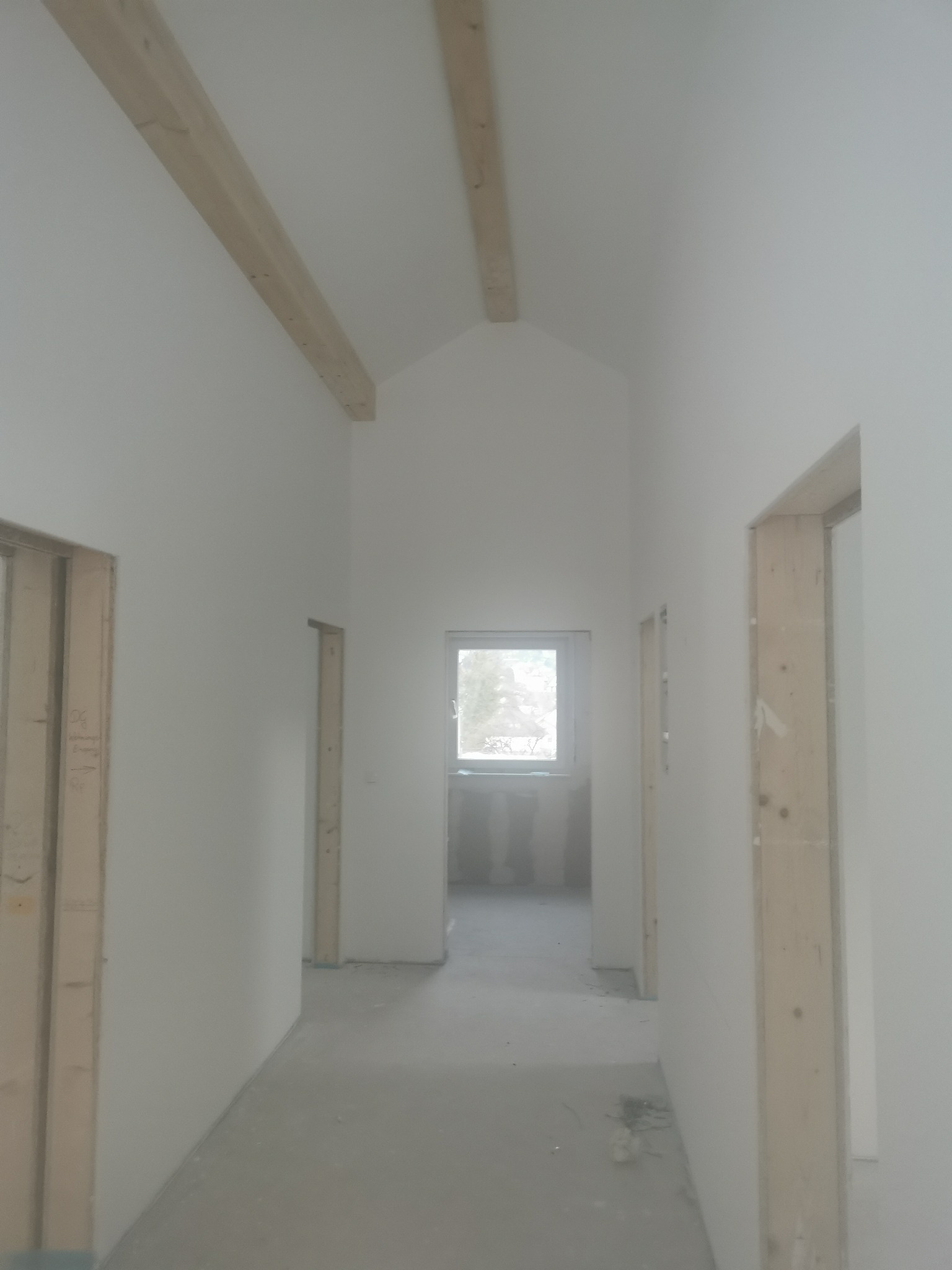 Wnętrze w trakcie wykończenia: białe ściany, drewniane belki stropowe, surowe ościeżnice. Widok na okno na końcu korytarza. Minimalistyczny, skandynawski styl.