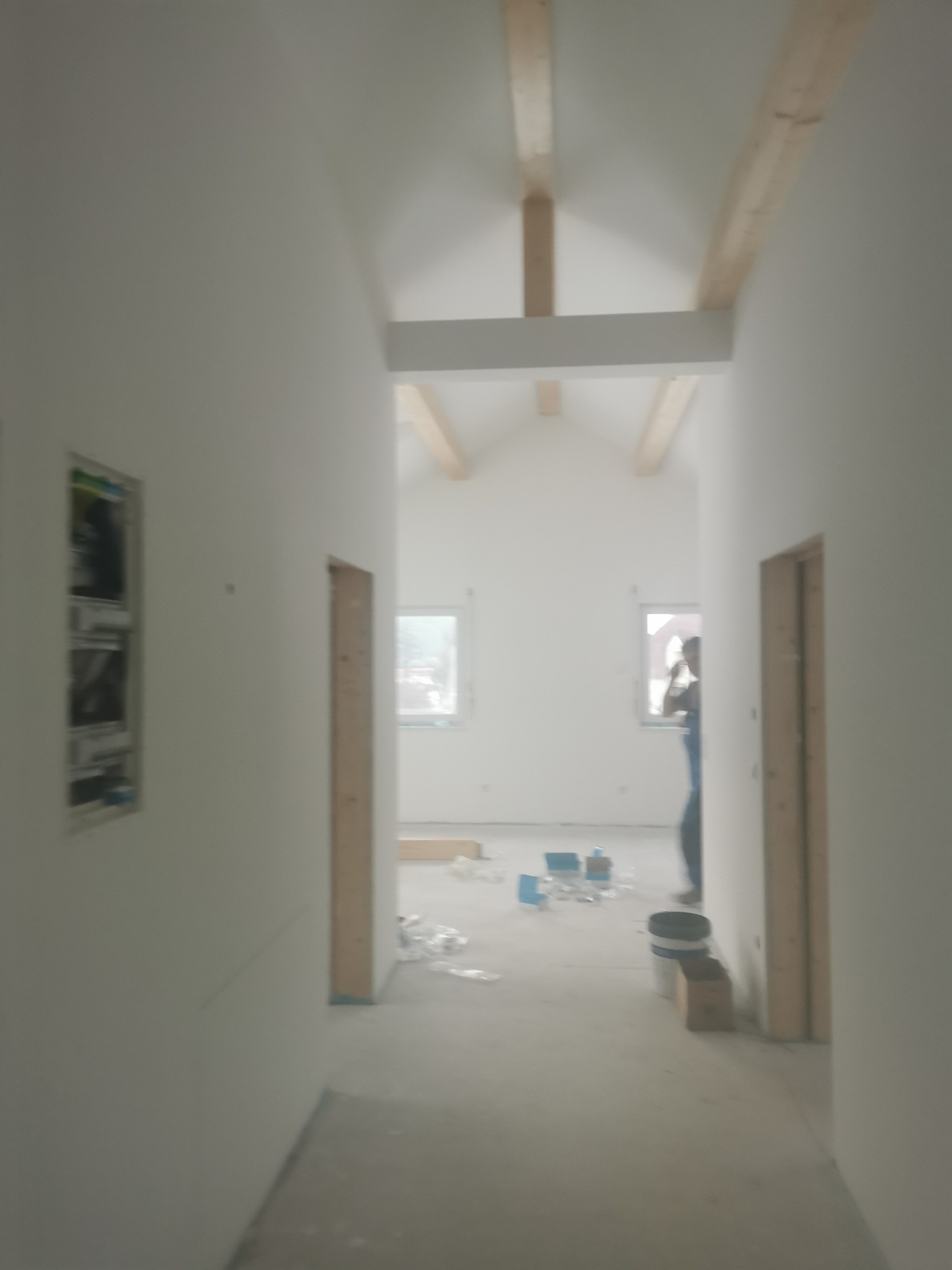Wnętrze domu w trakcie wykończenia: białe ściany, surowe drewniane belki stropowe, widoczne otwory drzwiowe i okno z sylwetką osoby. Na podłodze resztki materiałów budowlanych.